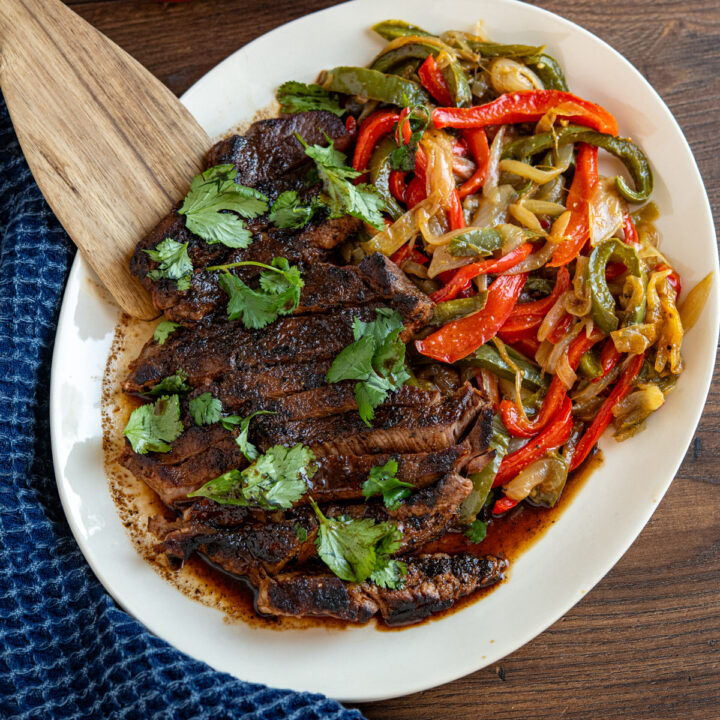 marinated steak on oval plate with sauteed peppers and onions, garnished with cilantro.