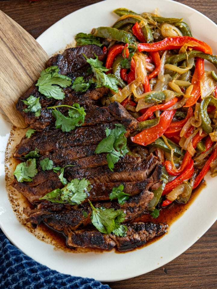 marinated steak on oval plate with sauteed peppers and onions, garnished with cilantro.
