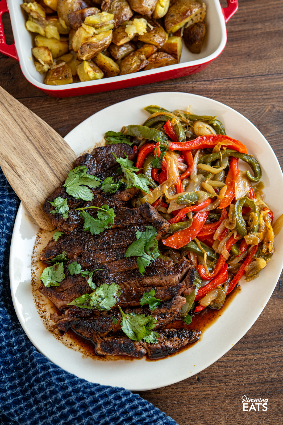Tender, flavour packed steak marinated in spices and finished with a sweet savory glaze for a simple yet delicious meal. Marinated Mouth Watering Steak on oval cream plate with sauteed onions and peppers,red dish with side of potatoes visible above.