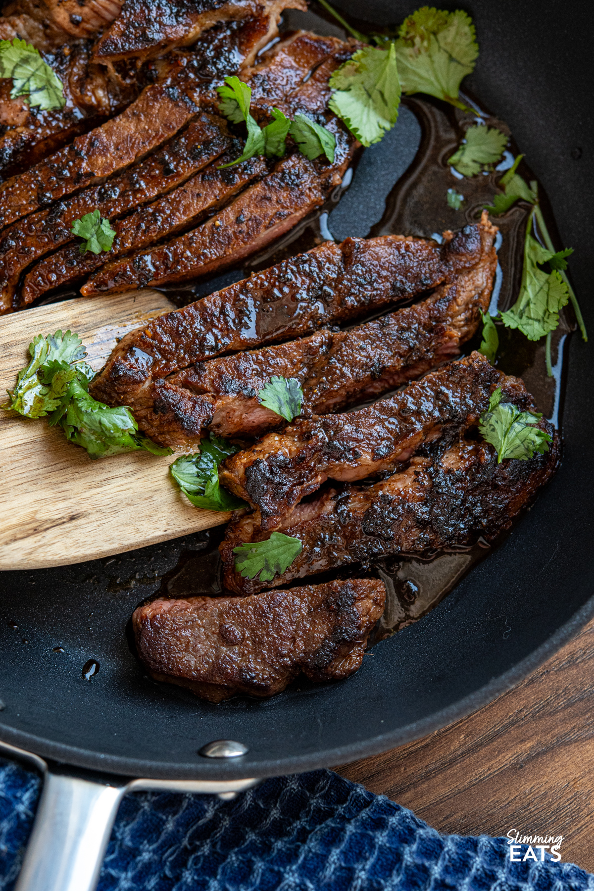 Tender, flavour packed steak marinated in spices and finished with a sweet savory glaze for a simple yet delicious meal. close up of Marinated Mouth Watering Steak in a non stick frying pan with wooden spatula and coriander garnish