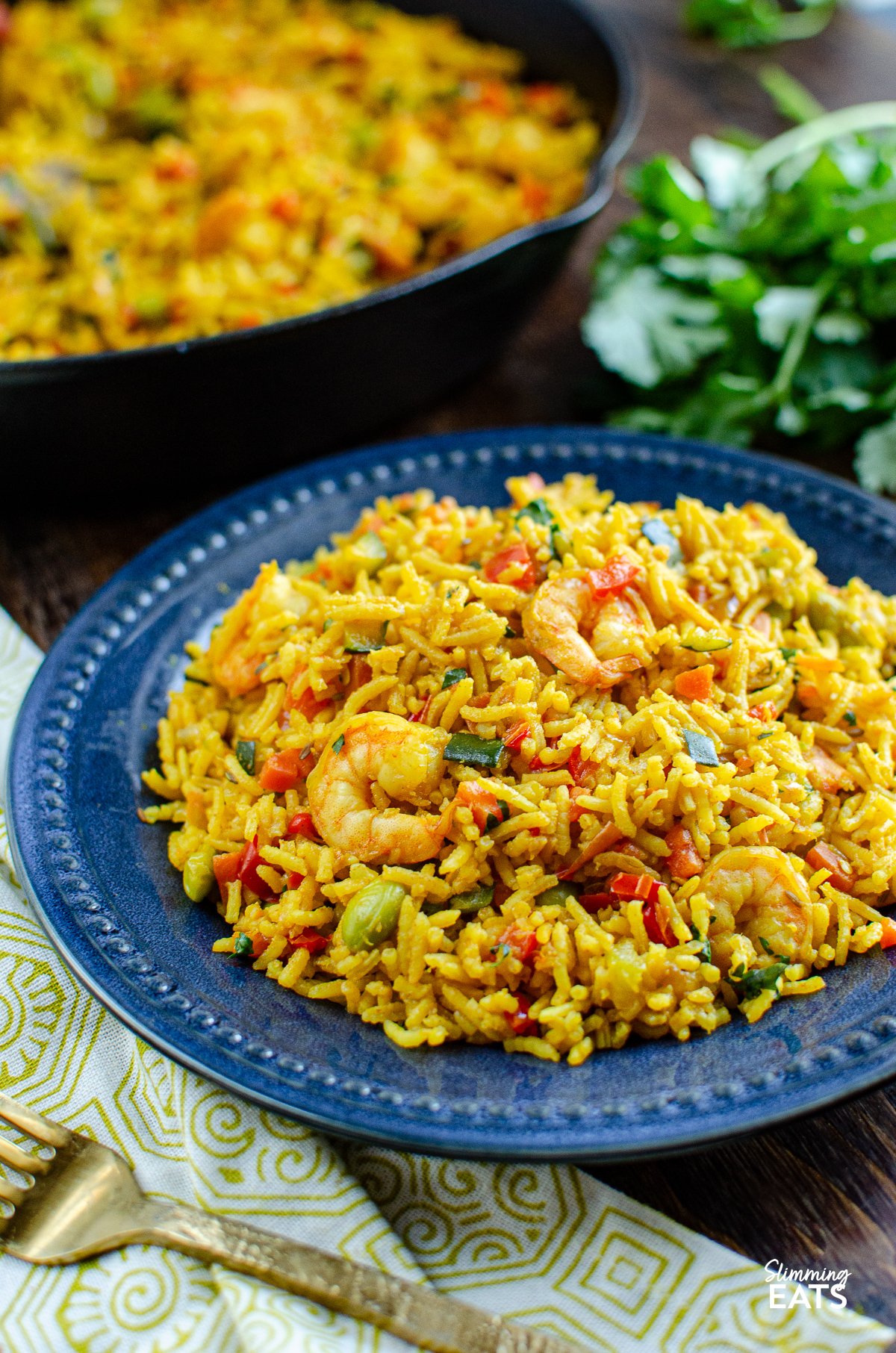 One Pot Prawn and Vegetable Pilaf on a navy blue plate with skillet and coriander in background