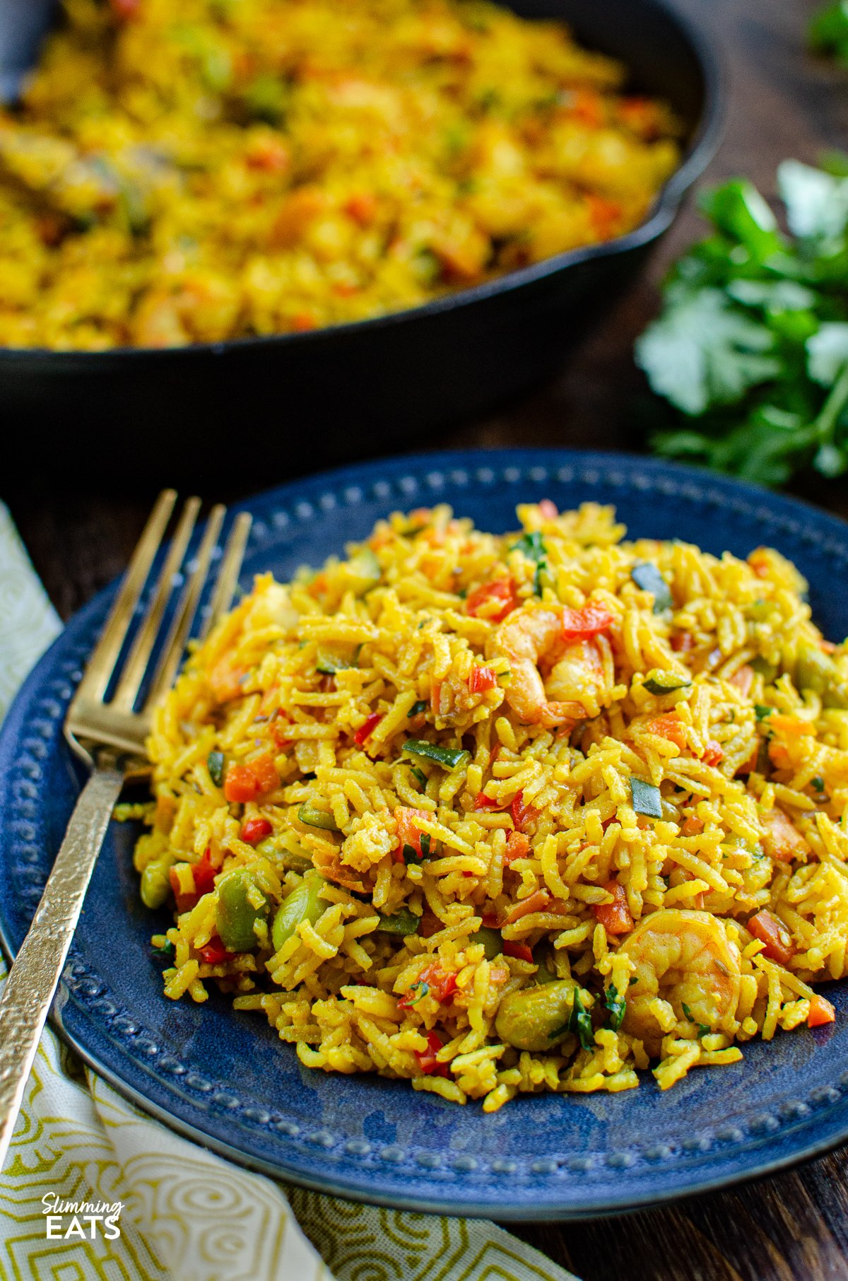 One Pot Prawn and Vegetable Pilaf on navy plate with folk fork, skillet and coriander in background