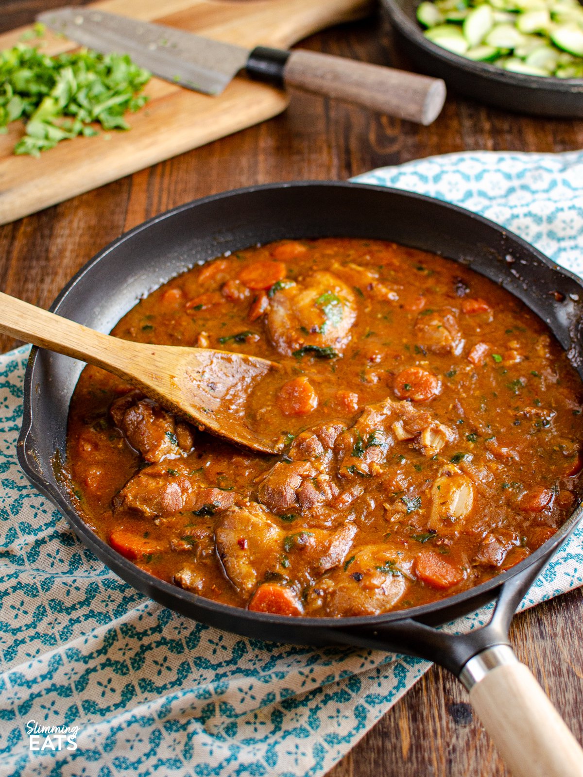 spicy ginger chicken in a black frying pan with wooden handle, wooden spoon in pan and chopping boad with coriander in background