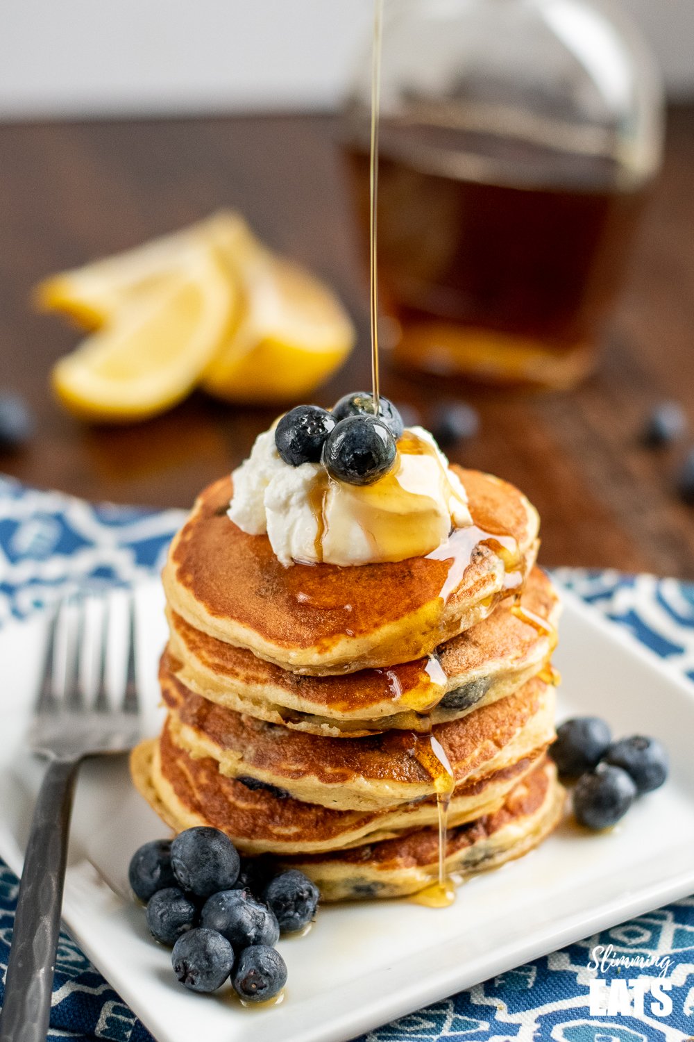 maple syrup being drizzled over lemon blueberry oat pancakes on a white plate