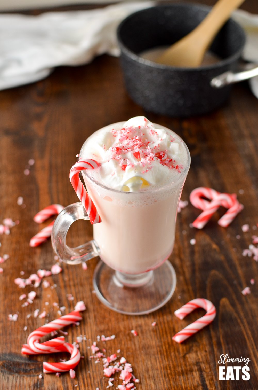 candy cane white hot chocolate in glass mug with saucepan in background and scattered broken candy canes
