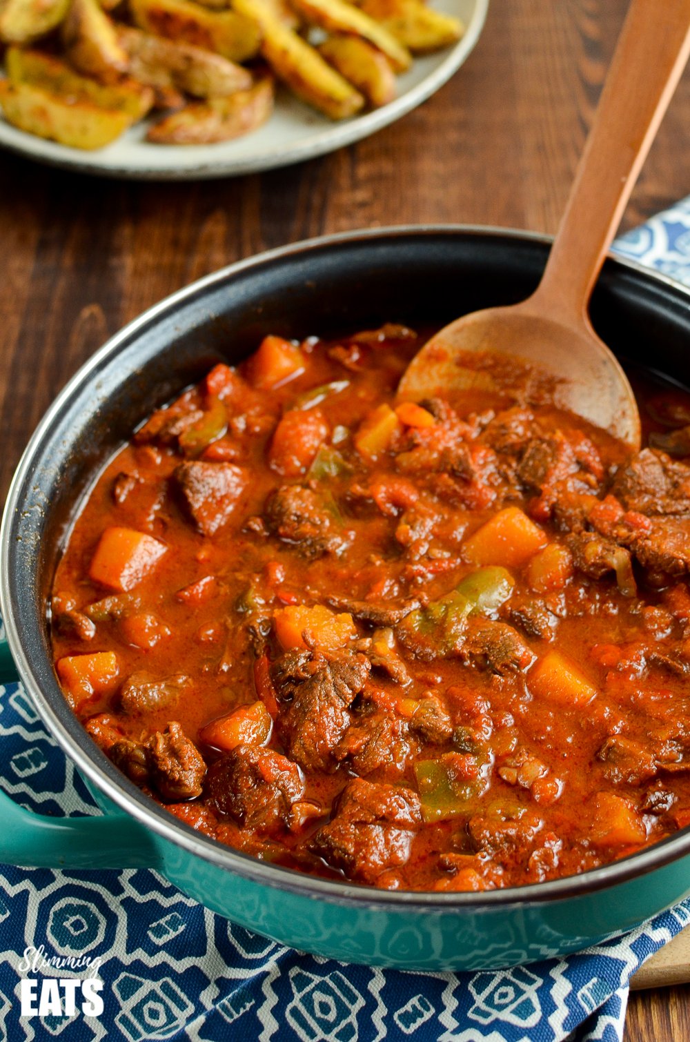 paprika beef in a pan with wooden spoon, potato wedges on a plate in background