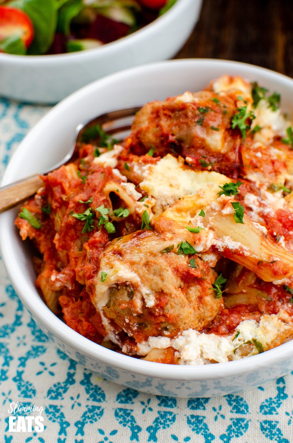 close up of slow cooker chicken meatballs pasta bake in white bowl with fork.