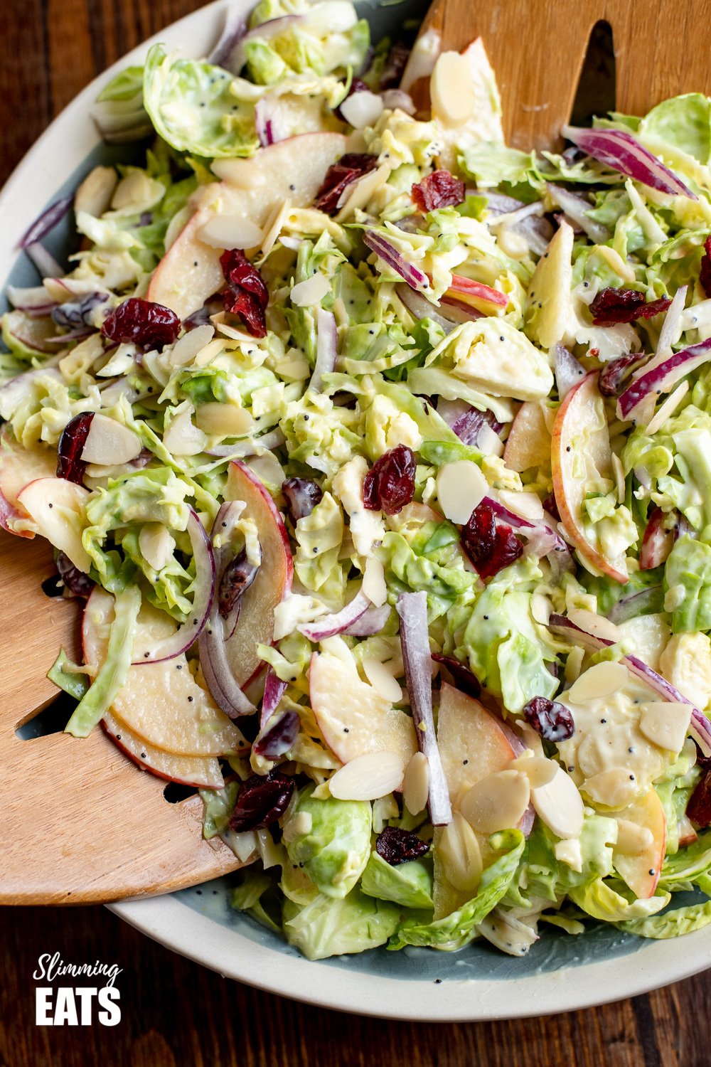 A delicious Healthy Brussels Sprouts Salad - with almond slices, crisp tart apple and sweet cranberries. Perfect on its own as a meal or as a side dish for your main Gluten Free, Vegetarian, Slimming and Weight Watchers friendly close up of tossed Brussel Sprouts salad in bowl with wooden salad tongs