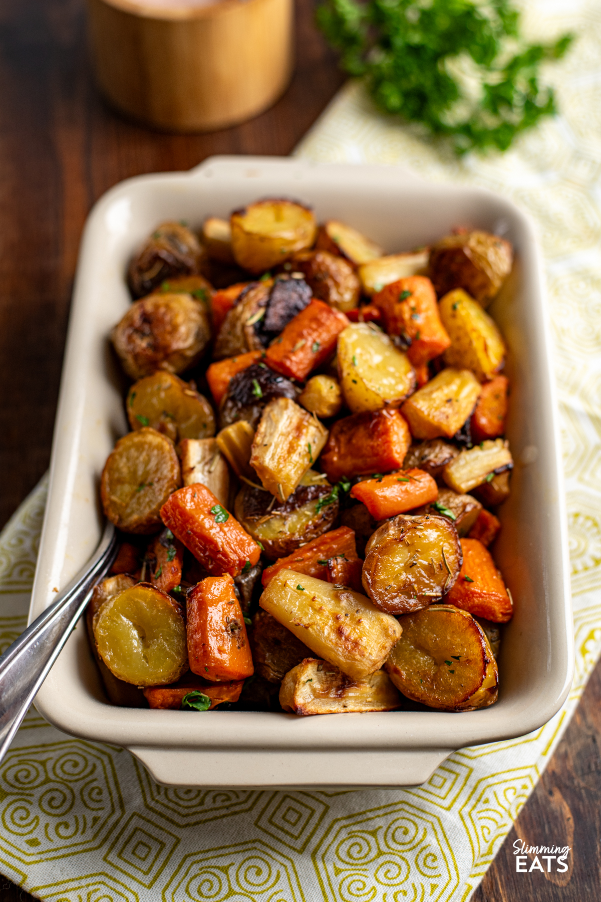 Rosemary Roasted Potatoes, Parsnips, Carrots and Onion in a dish on a pale olive green and white towel with parsley and salt canister in background