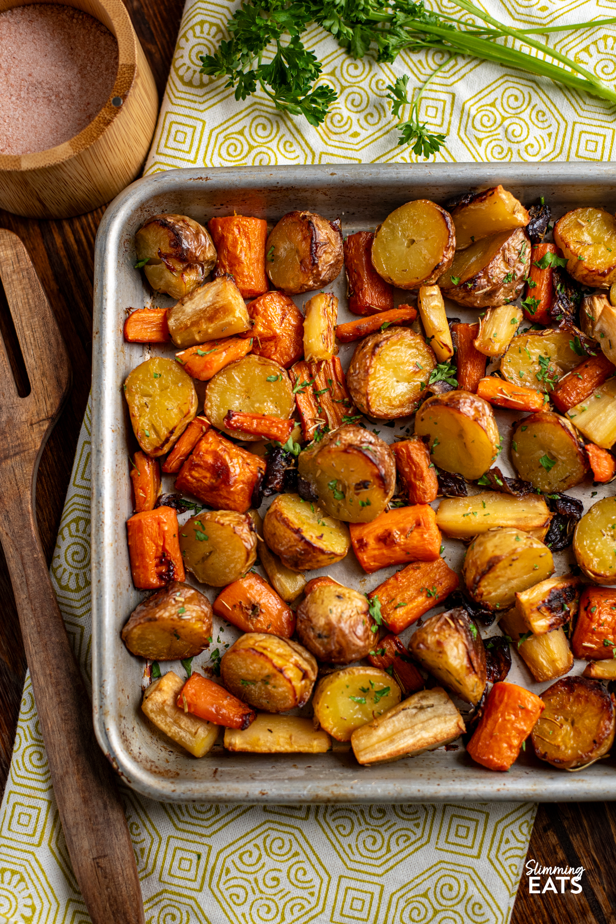 Rosemary Roasted Potatoes, Parsnips, Carrots and Onion on a baking tray next to a wooden spatula on a olive green and white towel