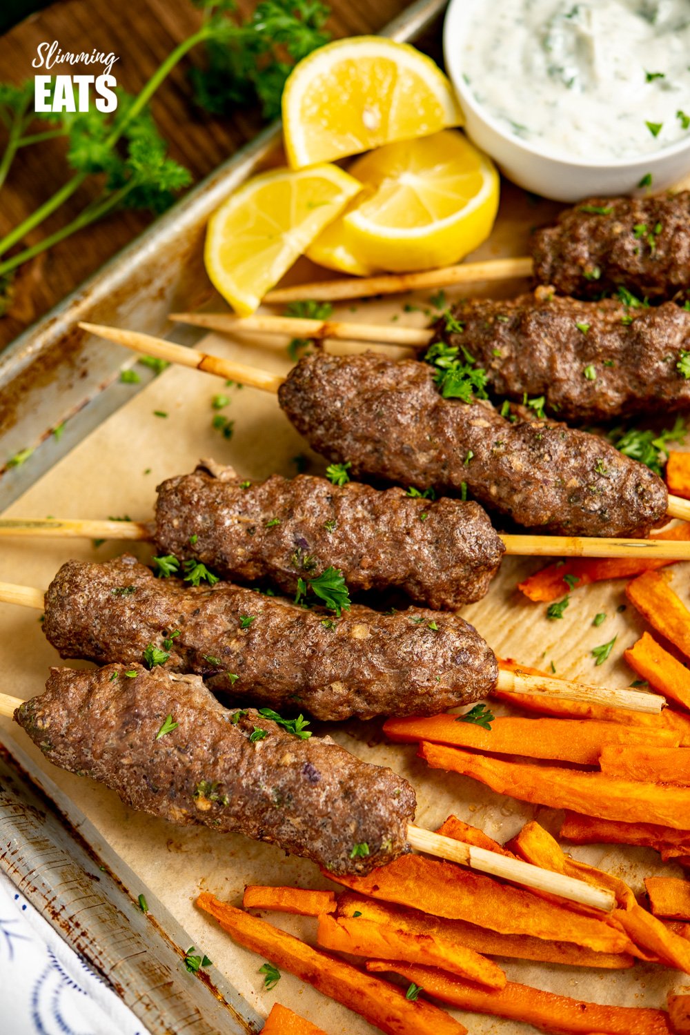 Yummy Beef Kofta Tray Bake - authentic Kofta kebabs with sweet potato fries and delicious roasted vegetables all finished off with a garlic yoghurt sauce. Gluten Free, Slimming Eats and Weight Watchers friendly close up of Beef Kofta Tray Bake on baking tray with white bowl of garlic yoghurt sauce and lemon slices