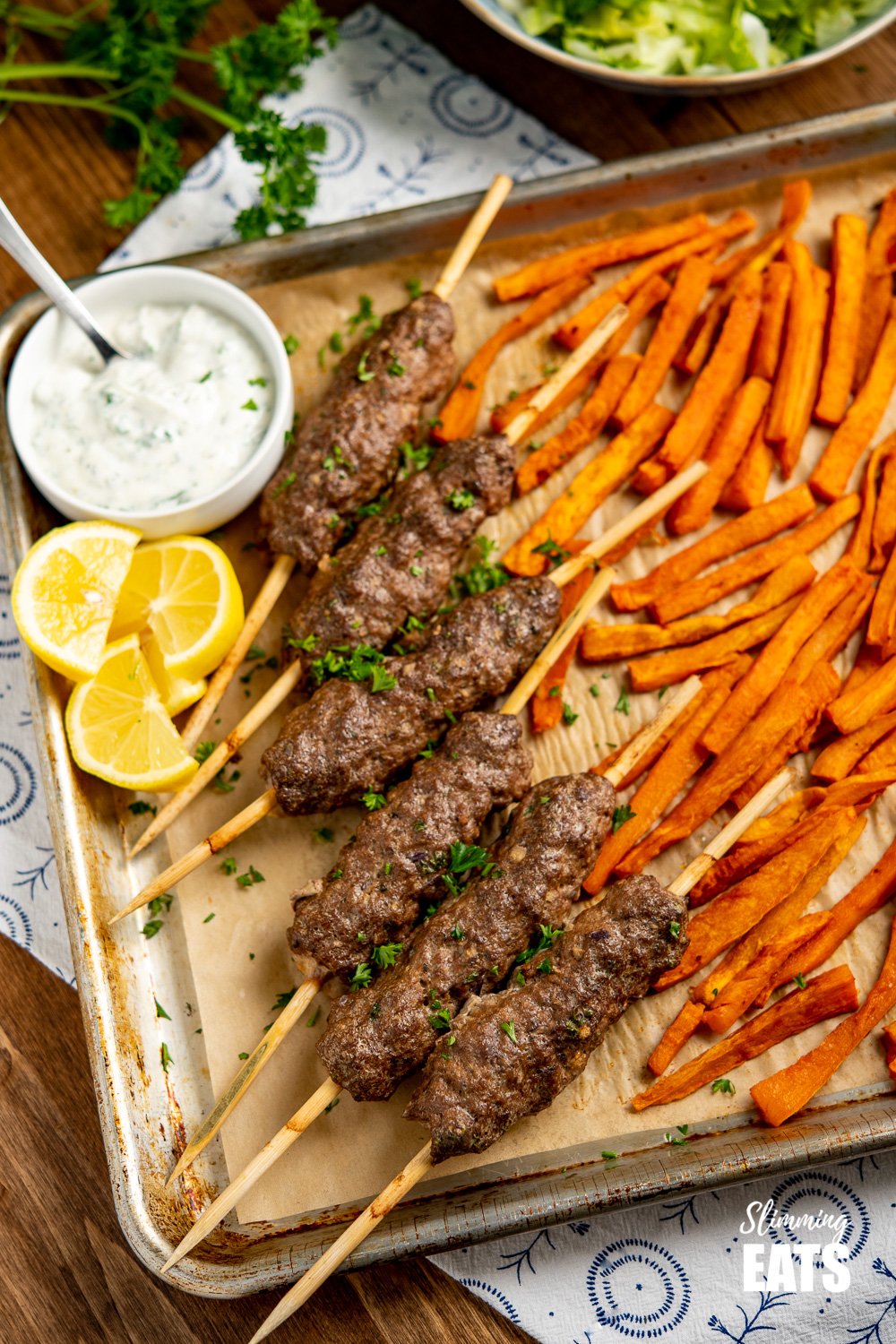 Beef Kofta Tray Bake with skewers or kofta, sweet potato fries, garlic yoghurt sauce and lemon slices on wooden board.