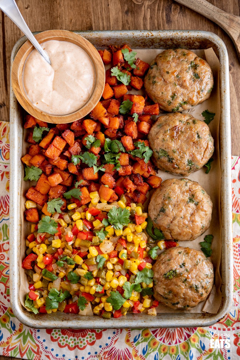 4 honey garlic chicken burgers on a baking tray with butternut squash, peppers, corn and dip