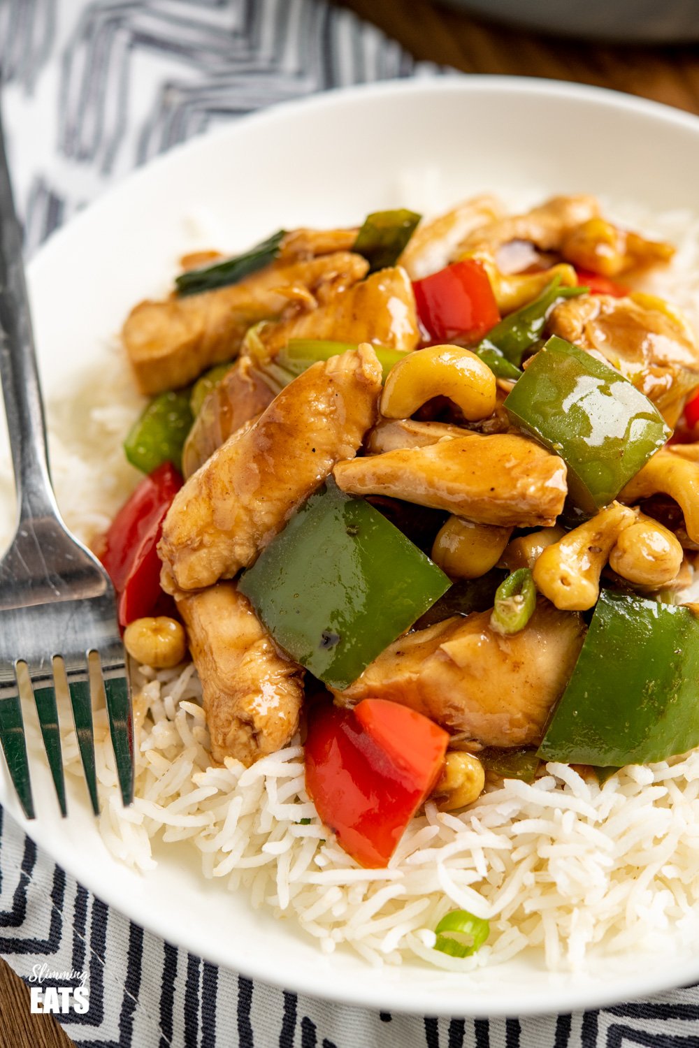 close up of Chinese cashew chicken on white shallow bowl with rice and black handled fork