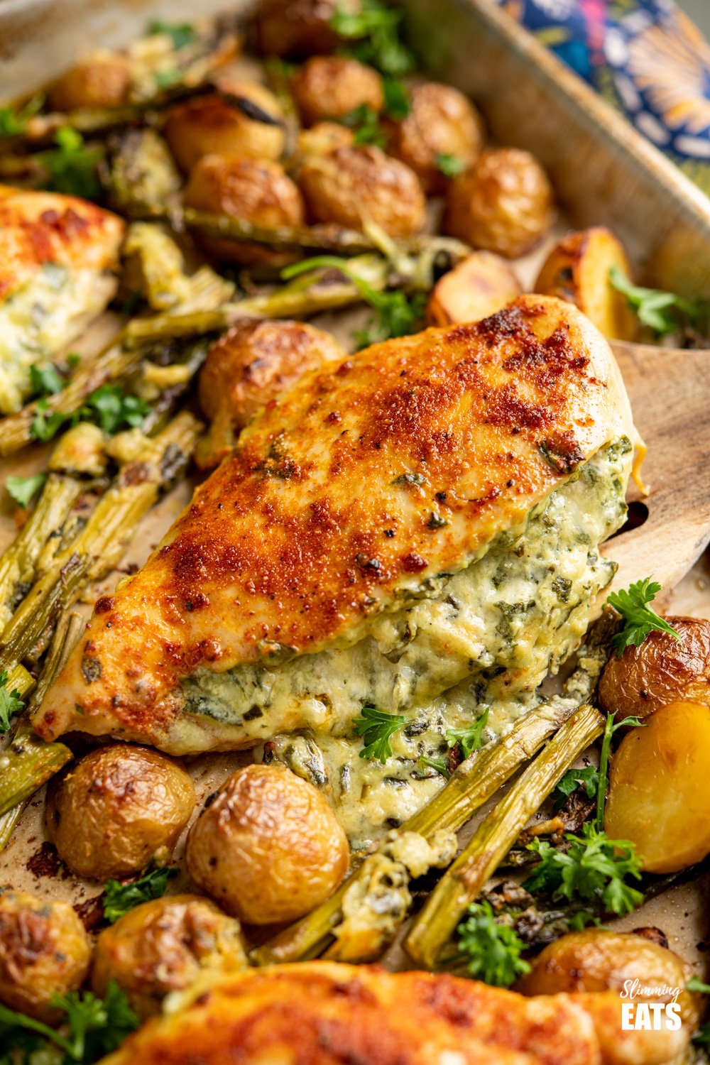 Creamy Cheesy Spinach Stuffed Chicken Tray Bake - Tender Juicy Chicken Breasts with a Creamy Parmesan Spinach Stuffing all baked on one tray with Baby Yukon Gold Potatoes and Asparagus. Gluten-Free, Slimming World and Weight Watchers friendly spatula lifting Creamy Cheesy Spinach Stuffed Chicken from baking tray