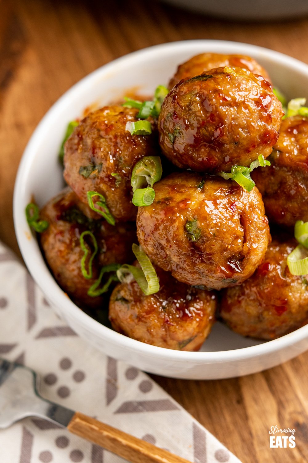 close up of sweet chilli meatballs in white bowl sprinkled with chopped spring onion.