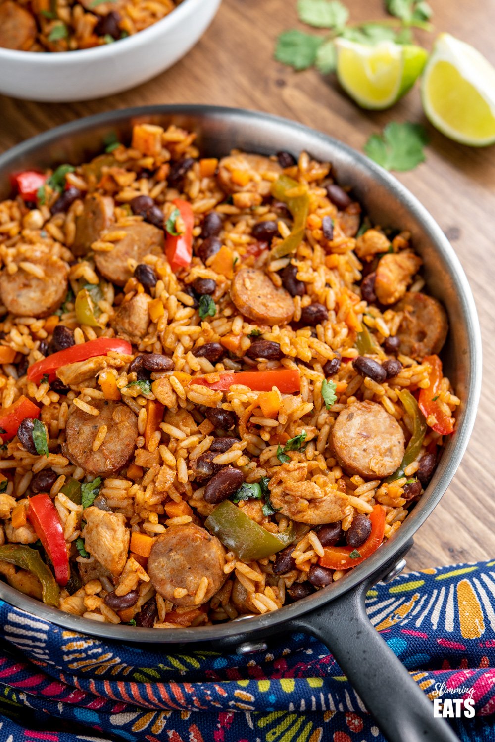 One Pot Cuban Style Rice (with Chicken and Sausage) - this delicious one-pot recipe with spicy seasonings, fresh vegetables and black beans is a perfect meal for any night of the week. Gluten Free, Dairy Free, Slimming Eats and Weight Watchers friendly One Pot Cuban Style Rice in frying pan with white filled bowl in background and lime slices