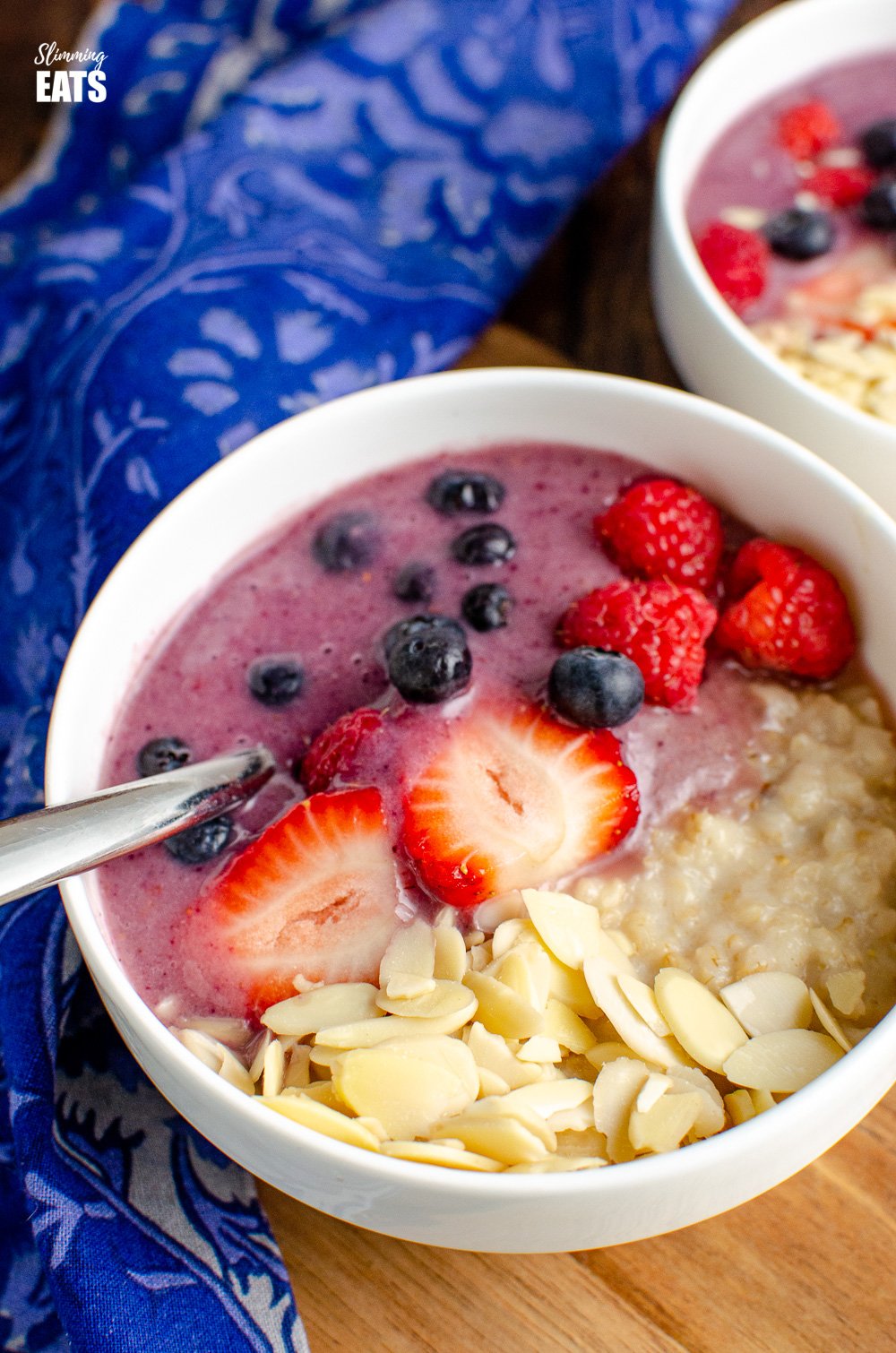 oatmeal smoothie bowl in white bowl with silver spoon on wooden board with blue patterned napkin to left.