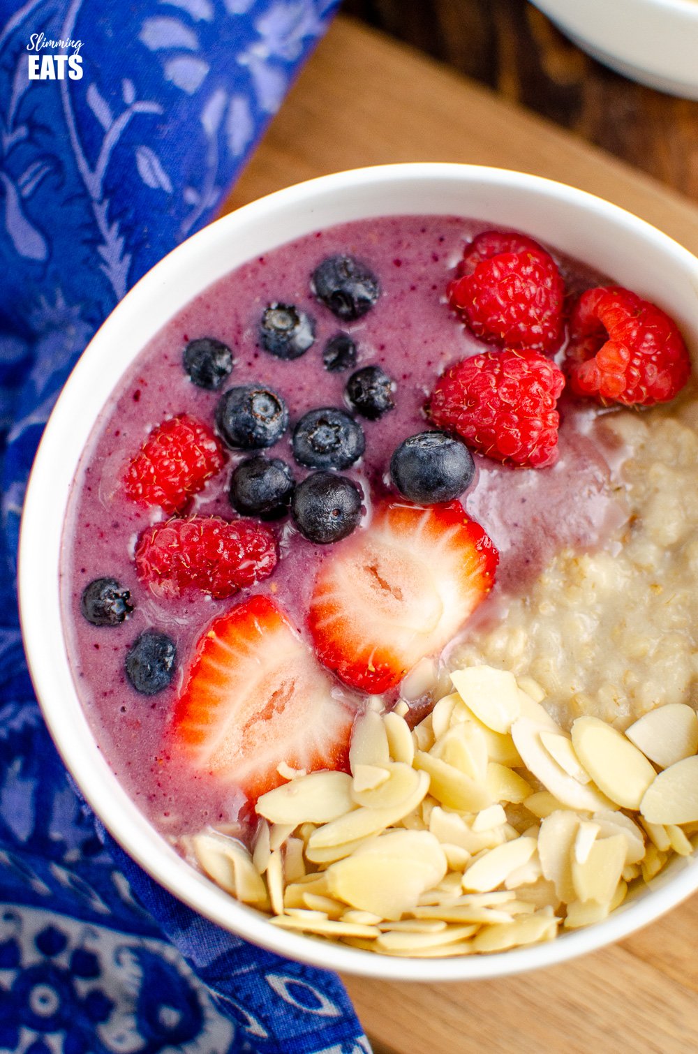Oatmeal Smoothie Bowl - a filling healthy breakfast combining a delicious fruit smoothie, fresh fruit, almonds and oatmeal. Dairy Free, Gluten Free, Vegan, Vegetarian, Slimming Eats and Weight Watchers friendly close up of oatmeal smoothie bowl in white bowl on wooden board with blue patterned napkin to left