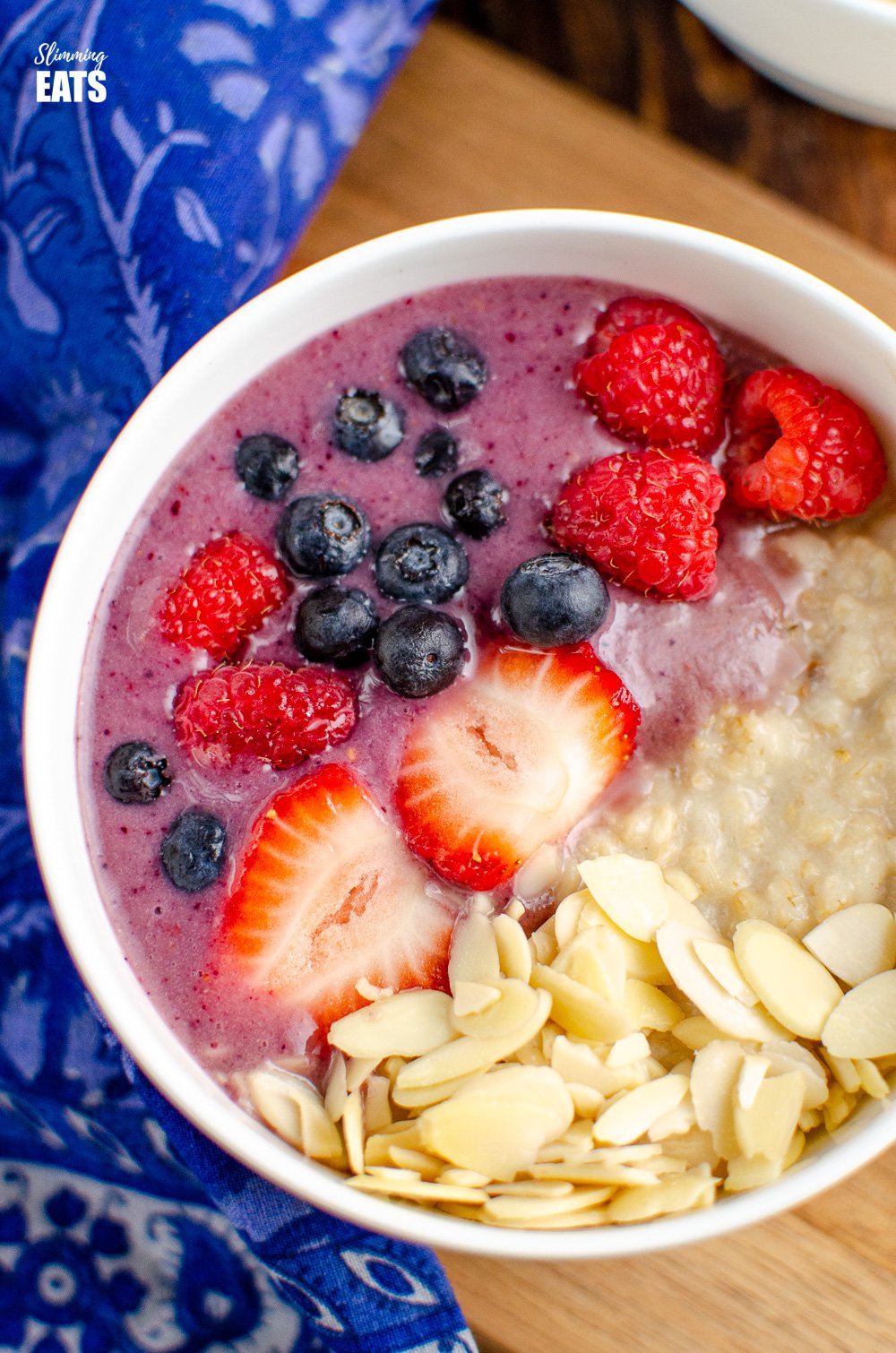 close up of oatmeal smoothie bowl in white bowl on wooden board with blue patterned napkin to left