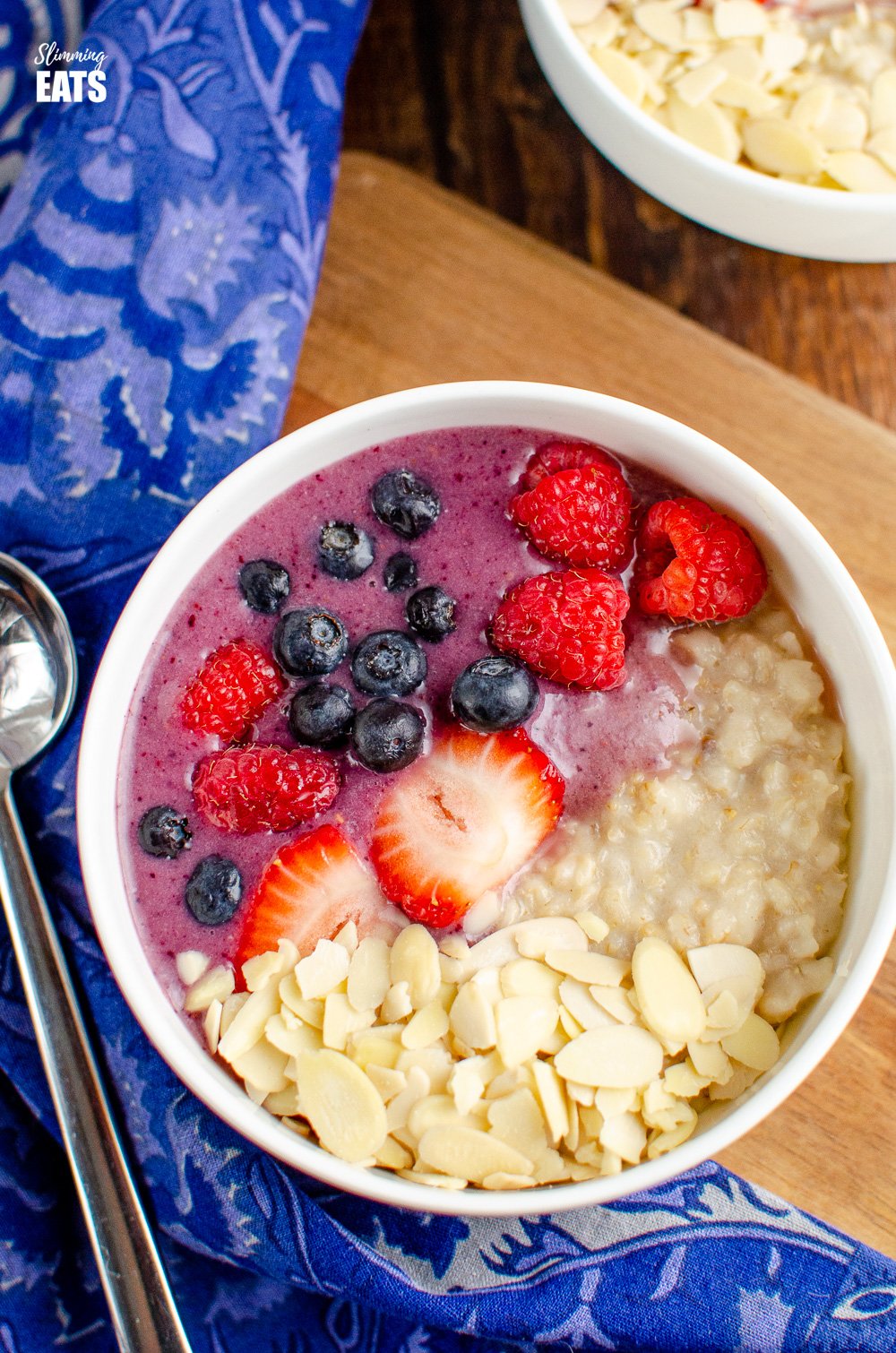 over top view of oatmeal smoothie bowl in white bowl on wooden board with blue patterned napkin to left with silver spoon