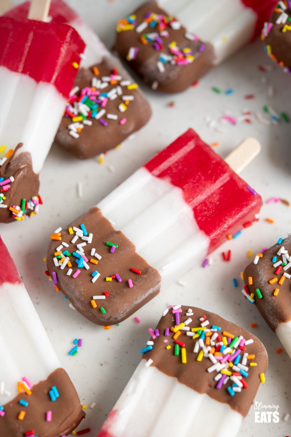 Homemade Fab Ice Lollies - a make at home version of the popular frozen UK treat, combining the flavours of coconut vanilla and strawberry water ice and dipped in milk chocolate and sprinkles. Slimming Eatsand Weight Watchers friendly close up of fab ice lolly on a white board with scattered ice lollies around it