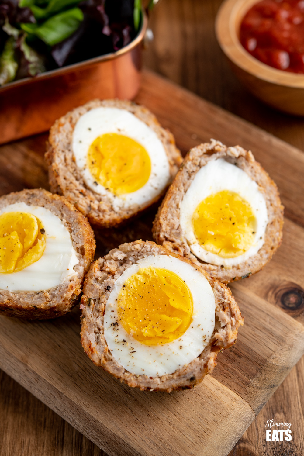 close up of Oven Baked Scotch Eggs sliced in half on a wooden board with salad and relish