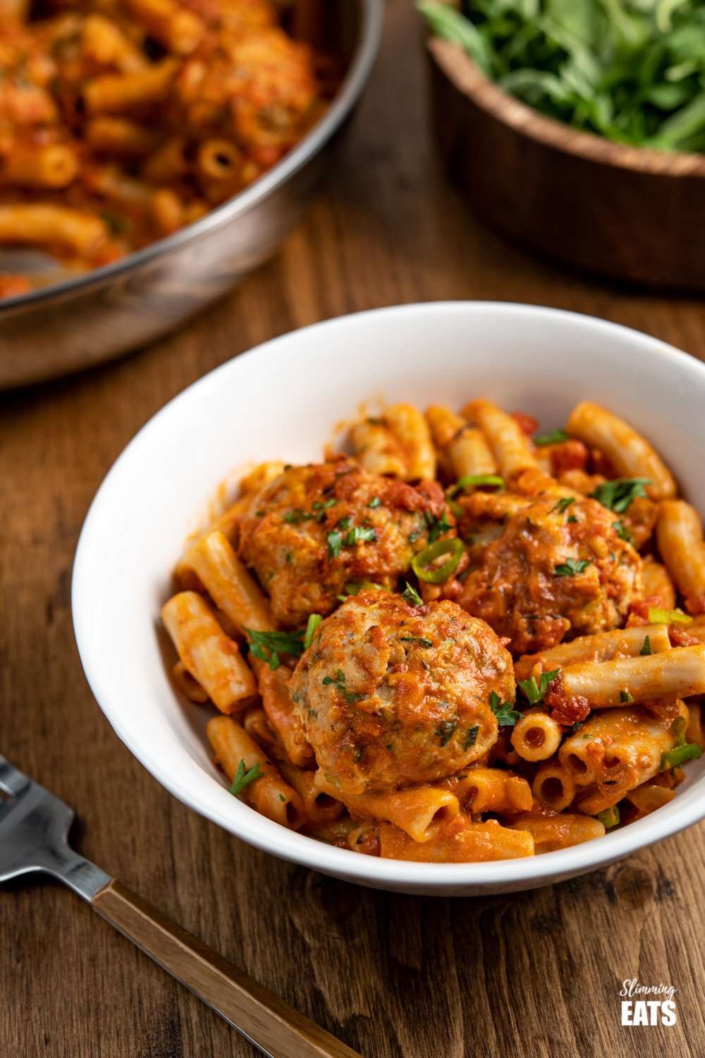 white bowl with creamy cajun meatball pasta and wooden bamboo bowl of arugula in background