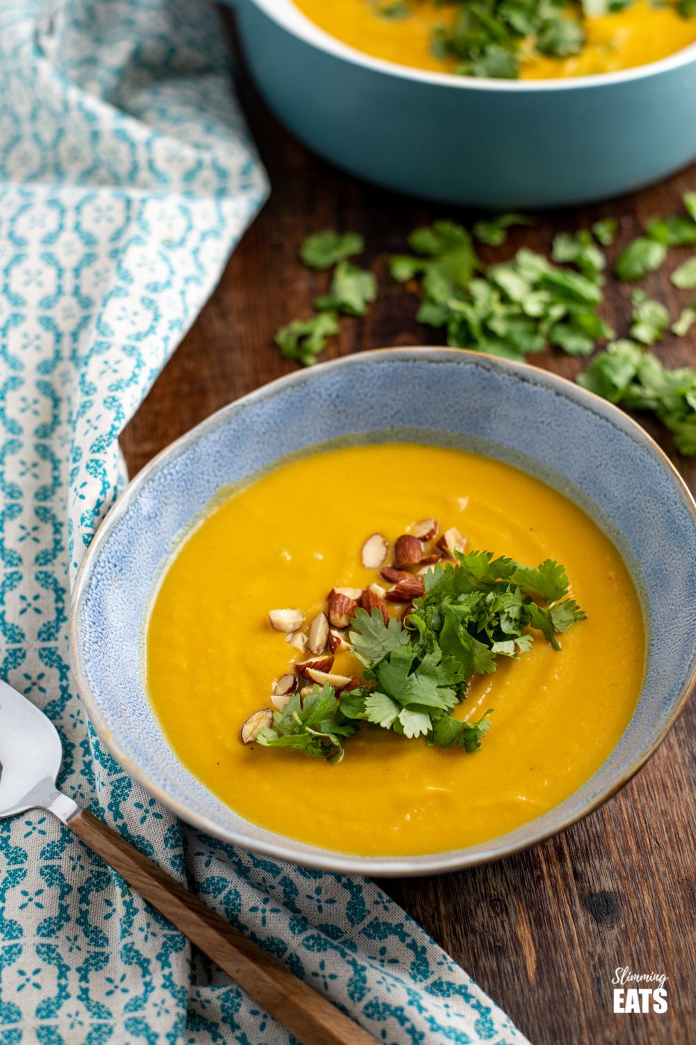 ginger carrot cauliflower soup in blue bowl topped with coriander and roughly chopped almonds on wooden board with scatter coriander