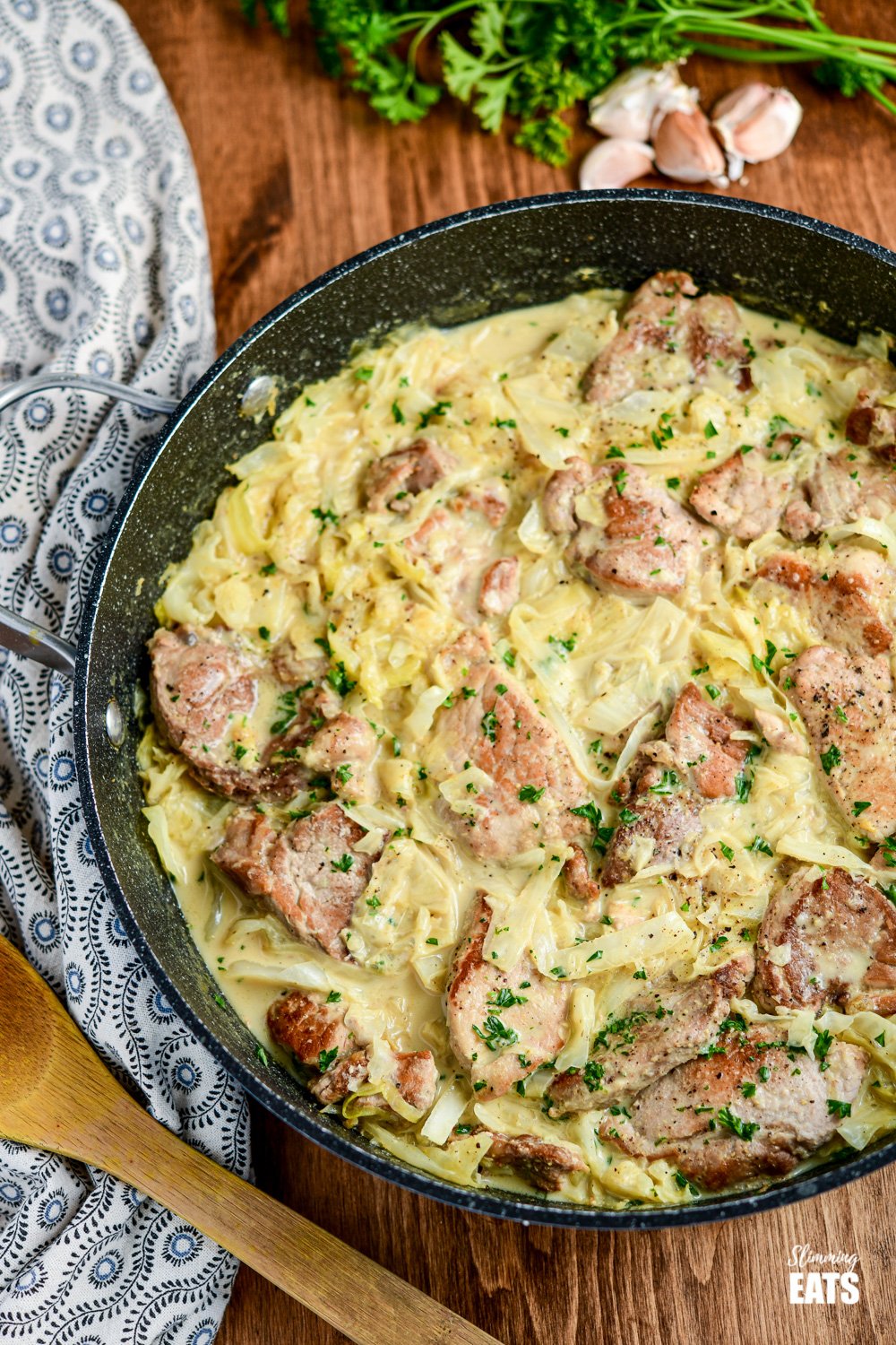 Creamy Garlic Pork with Cabbage in pan with wooden spoon and scattered garlic