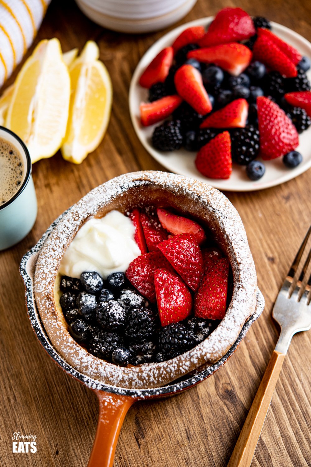 Dutch Baby in mini cast iron skillet with Berries and Yoghurt on a Wooden board with white bowl of berries and lemon wedge