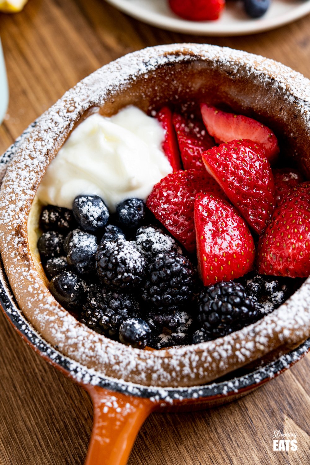 close up of Healthier Dutch Baby with Berries and Yoghurt in cast iron skillet dusted with icing sugar