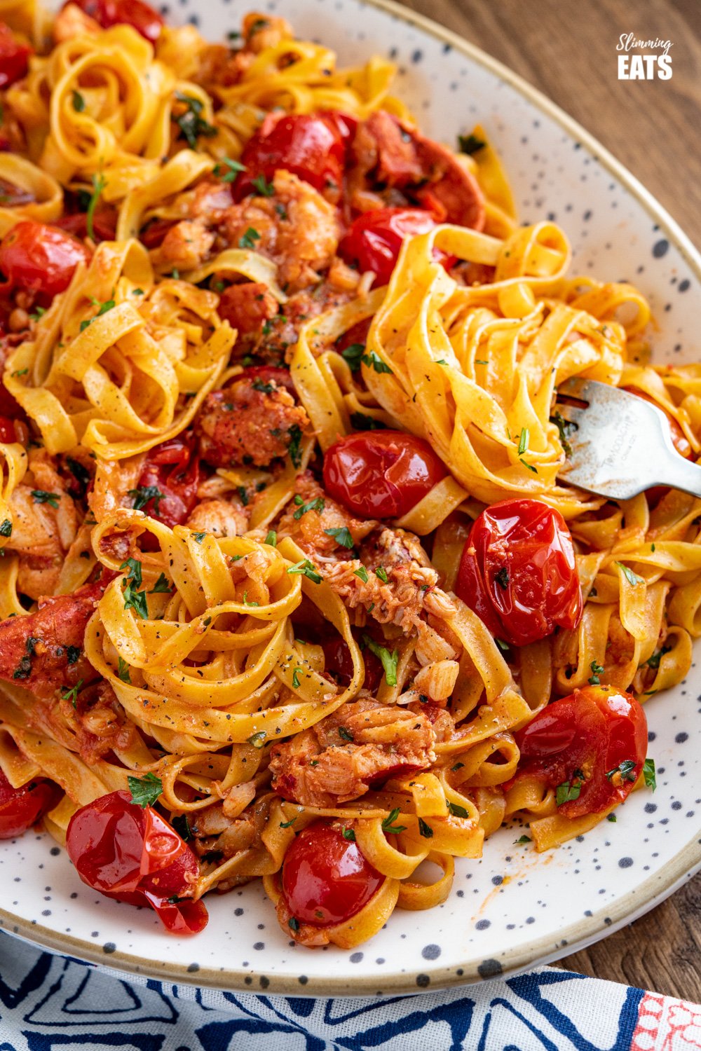 The most amazing fresh Lobster Pasta with the delicious sweet taste of fresh tomatoes and a lemon parsley buttery sauce. et taste of fresh tomatoes and a lemony garlic buttery sauce. Slimming Eats and Weight Watchers friendly fork spiralling up tagliatelle from speckled oval plate filled with lobster pasta