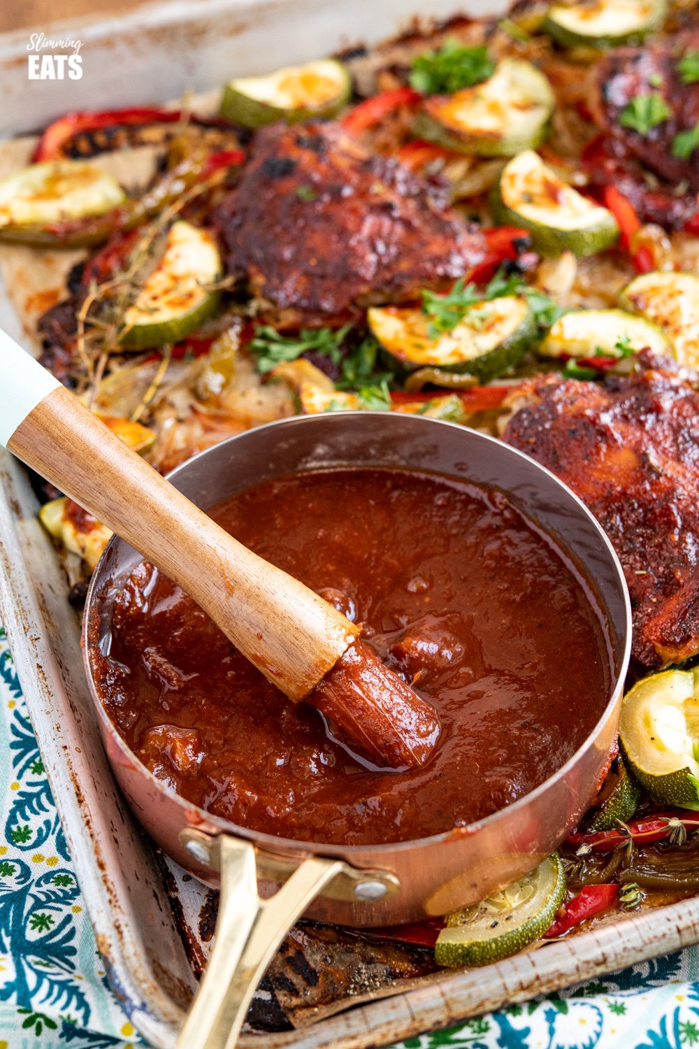 close up of small saucepan with basting brush in peach barbecue sauce on baking tray with chicken and veggies 