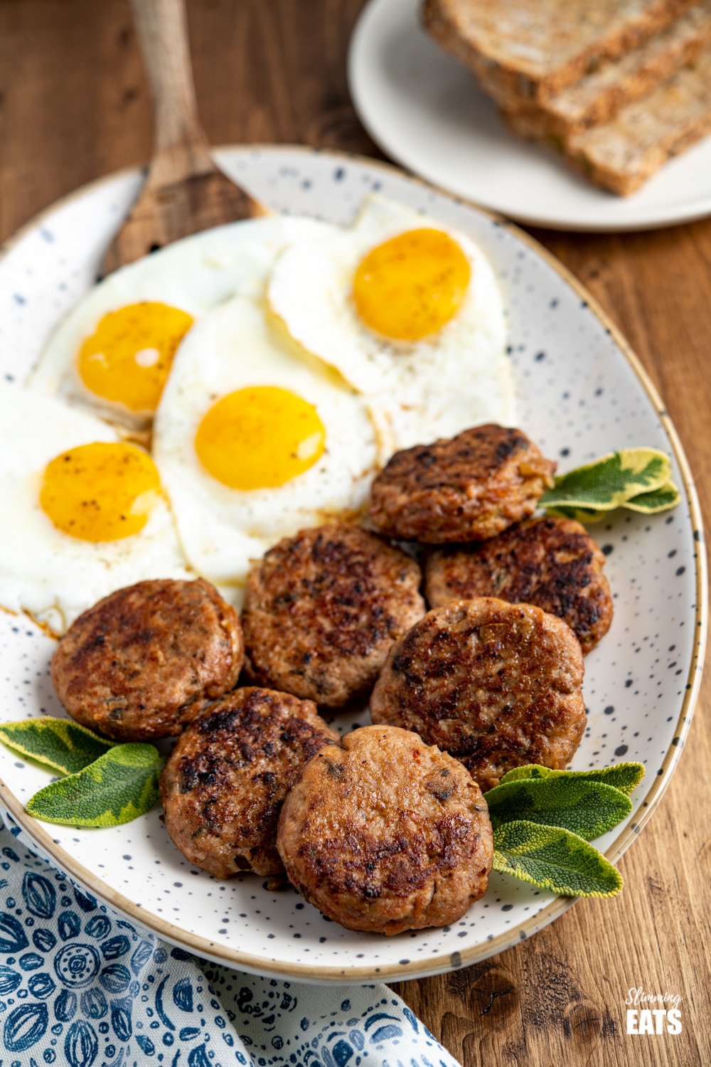 oval speckled plate filled with homemade turkey breakfast sausage patties and fried eggs, plate of wholemeal bread in the background