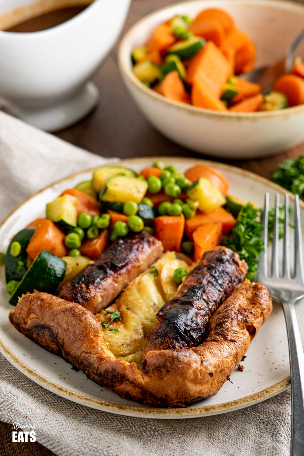 toad in the hole with vegetables on white and beige plate with fork and bowl of vegetables in background