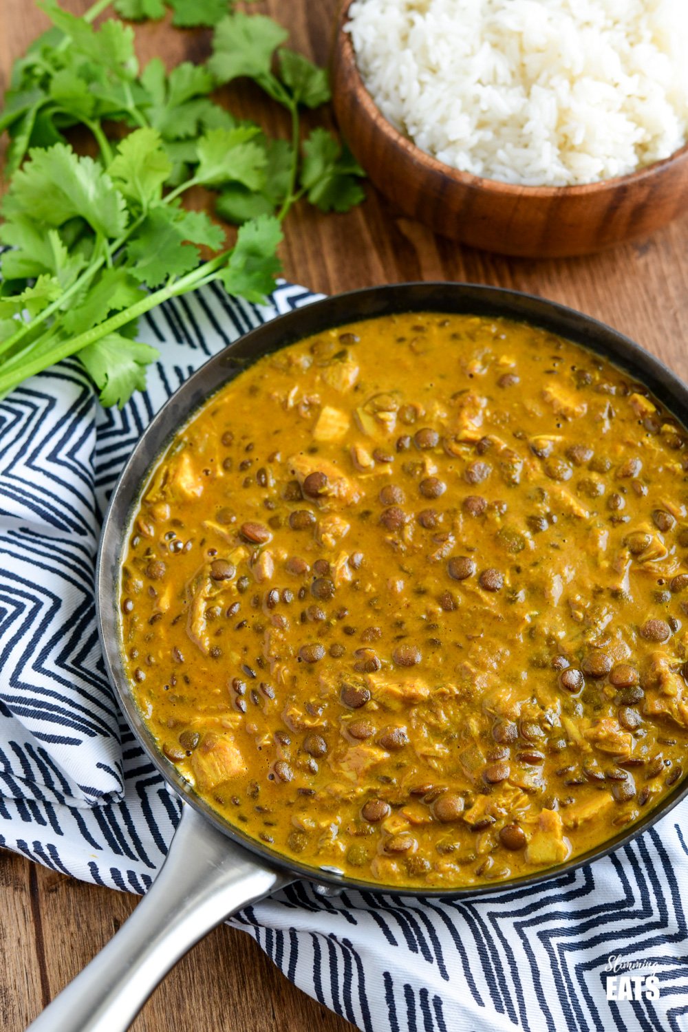 coconut chicken and lentil curry in a frying pan with a bamboo bowl filled with basmati rice and fresh coriander to the side