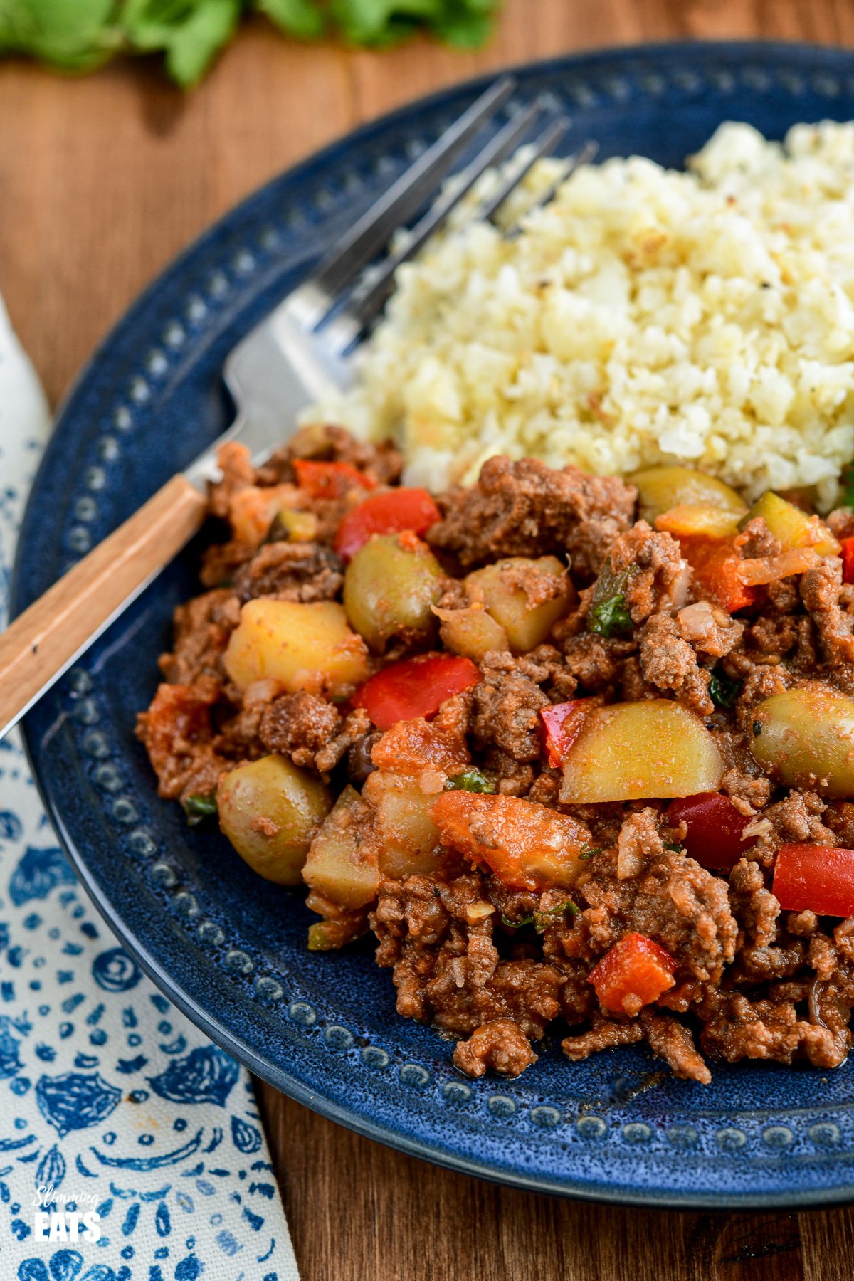 Cuban Beef Picadillo - a delicious hearty ground beef Cuban inspired recipe with flavoursome spices, potatoes, vegetables, olives and sultanas. #Cuban #picadillo #olives #beef #slimmingeats #weightwatchers #glutenfree #dairyfree close up of Cuban Beef Picadillo with cauliflower rice on dark blue plate.