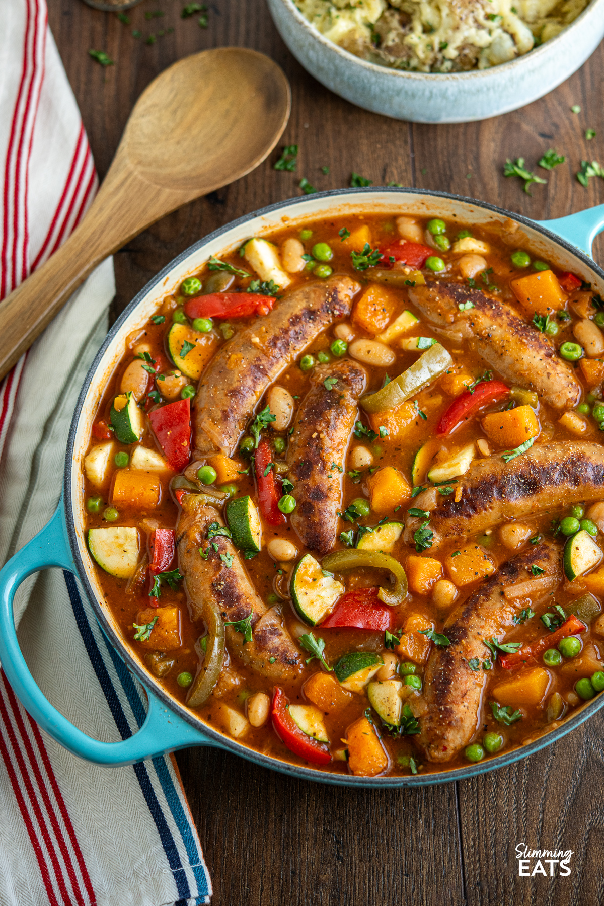 Sausage casserole with a variety of vegetables, presented in a turquoise cast iron pan, accompanied by a wooden spoon resting on the side along with bowl of mashed potatoes