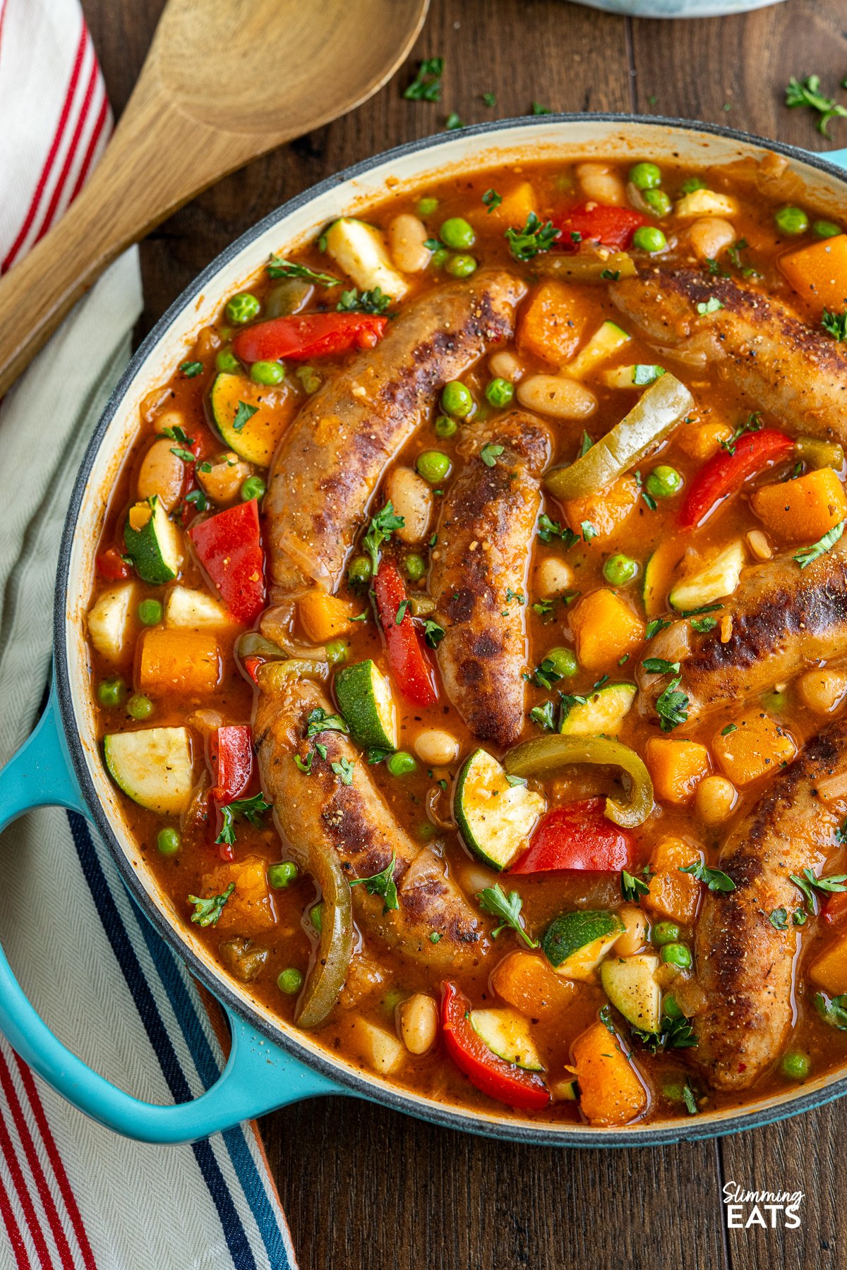 Sausage casserole with a variety of vegetables, presented in a turquoise cast iron pan, accompanied by a wooden spoon resting on the side