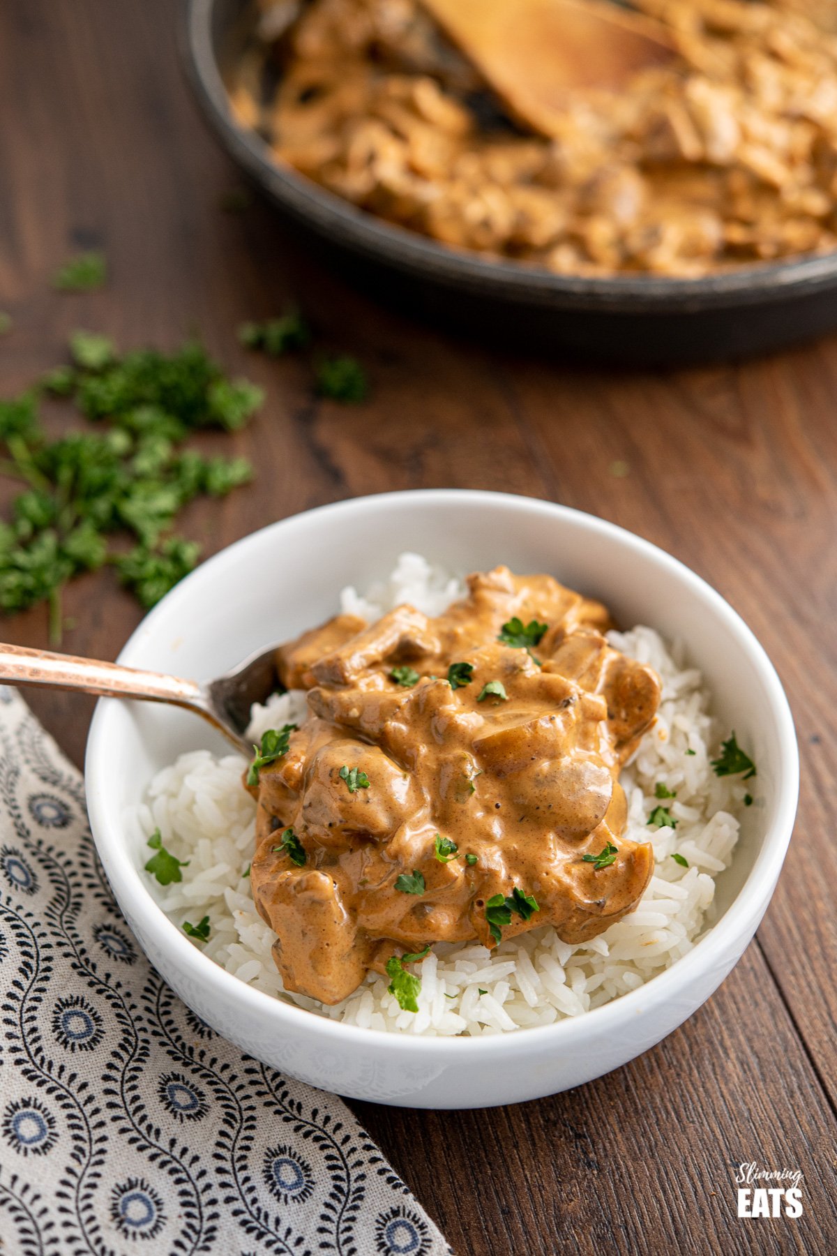 white bowl of Creamy Mushroom Stroganoff over rice on wooden board with frying pan and chopped parsley in background