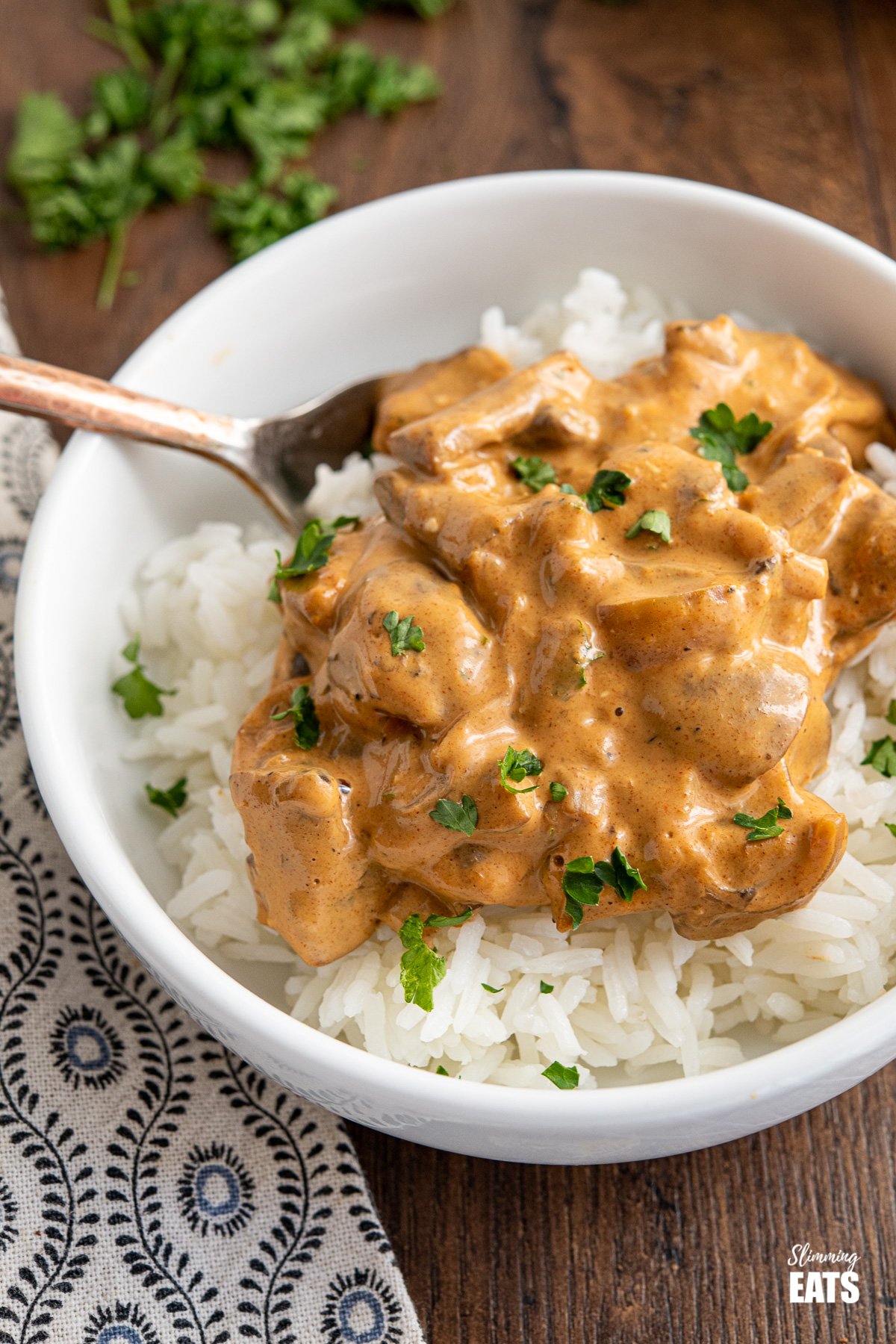 close up of white bowl of Creamy Mushroom Stroganoff over rice on wooden board with frying pan and chopped parsley in background