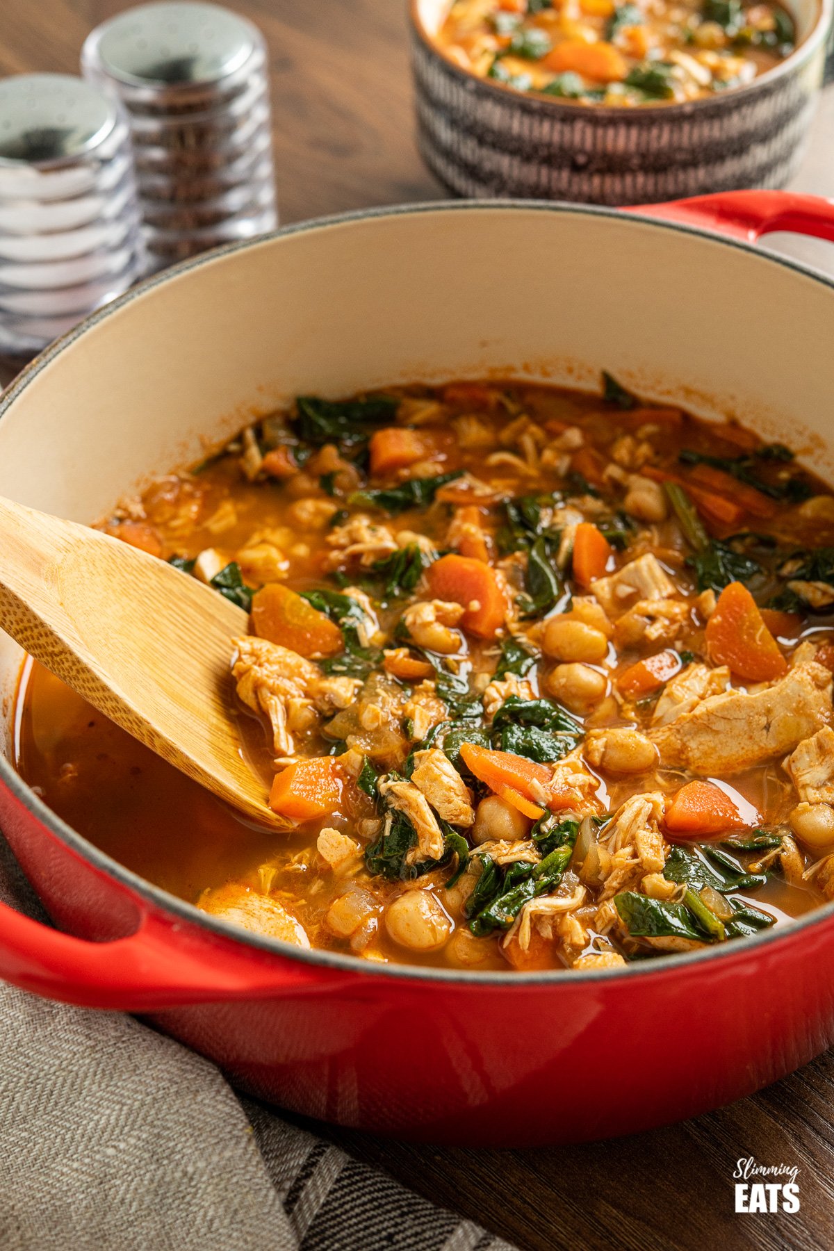 Chicken Chickpea and Spinach Soup in red cast iron port with wooden spoon on with cup of soup in backgrounders