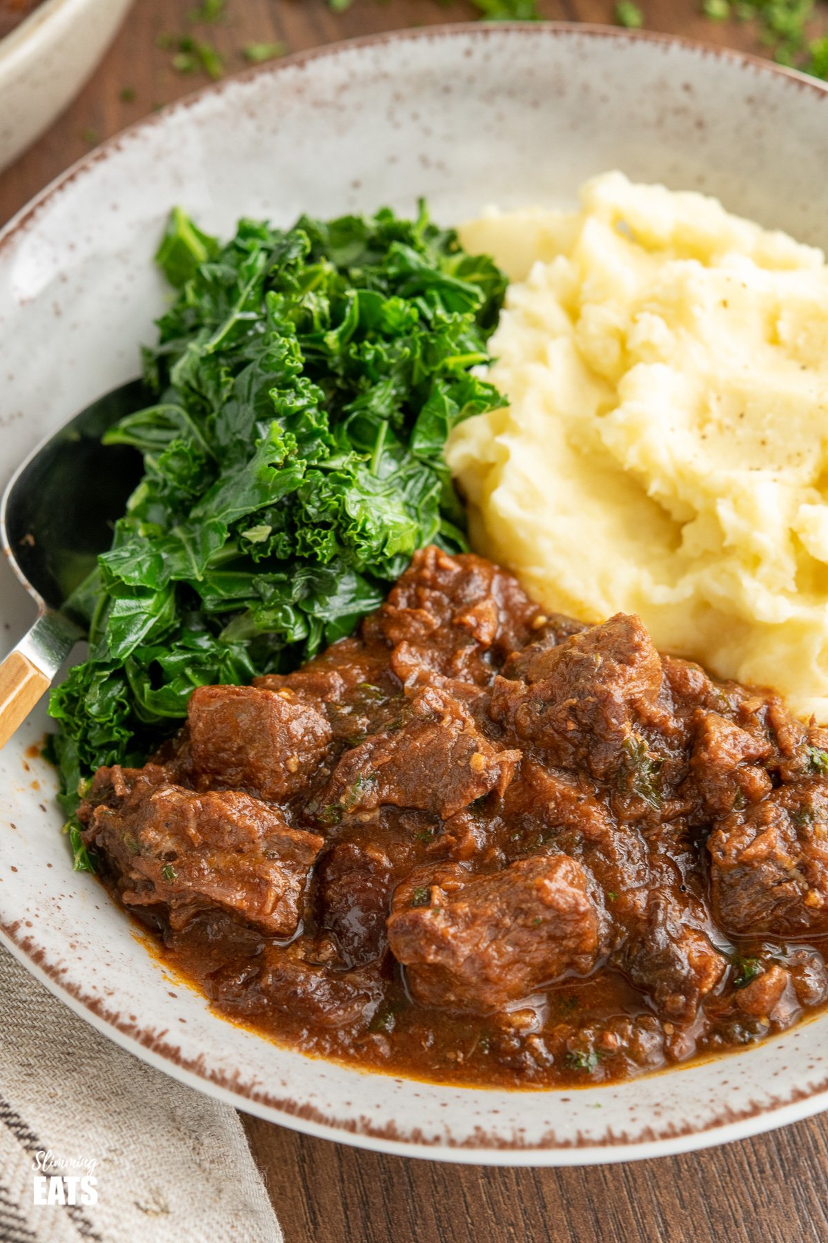 close up of beef and prune stew in beige bowl with mashed potatoes and kale