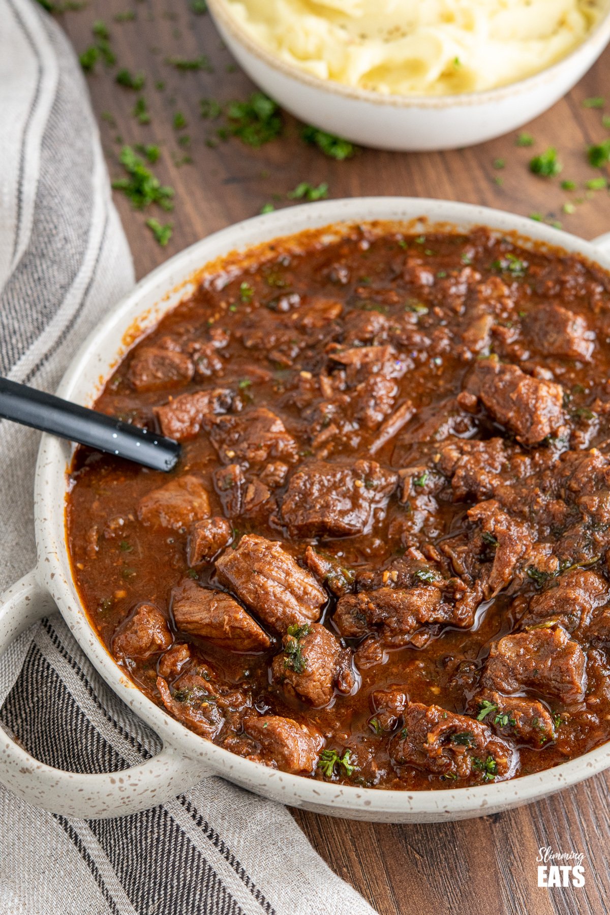 Beef and prune stew in brown grey serving dish with mashed potatoes in background