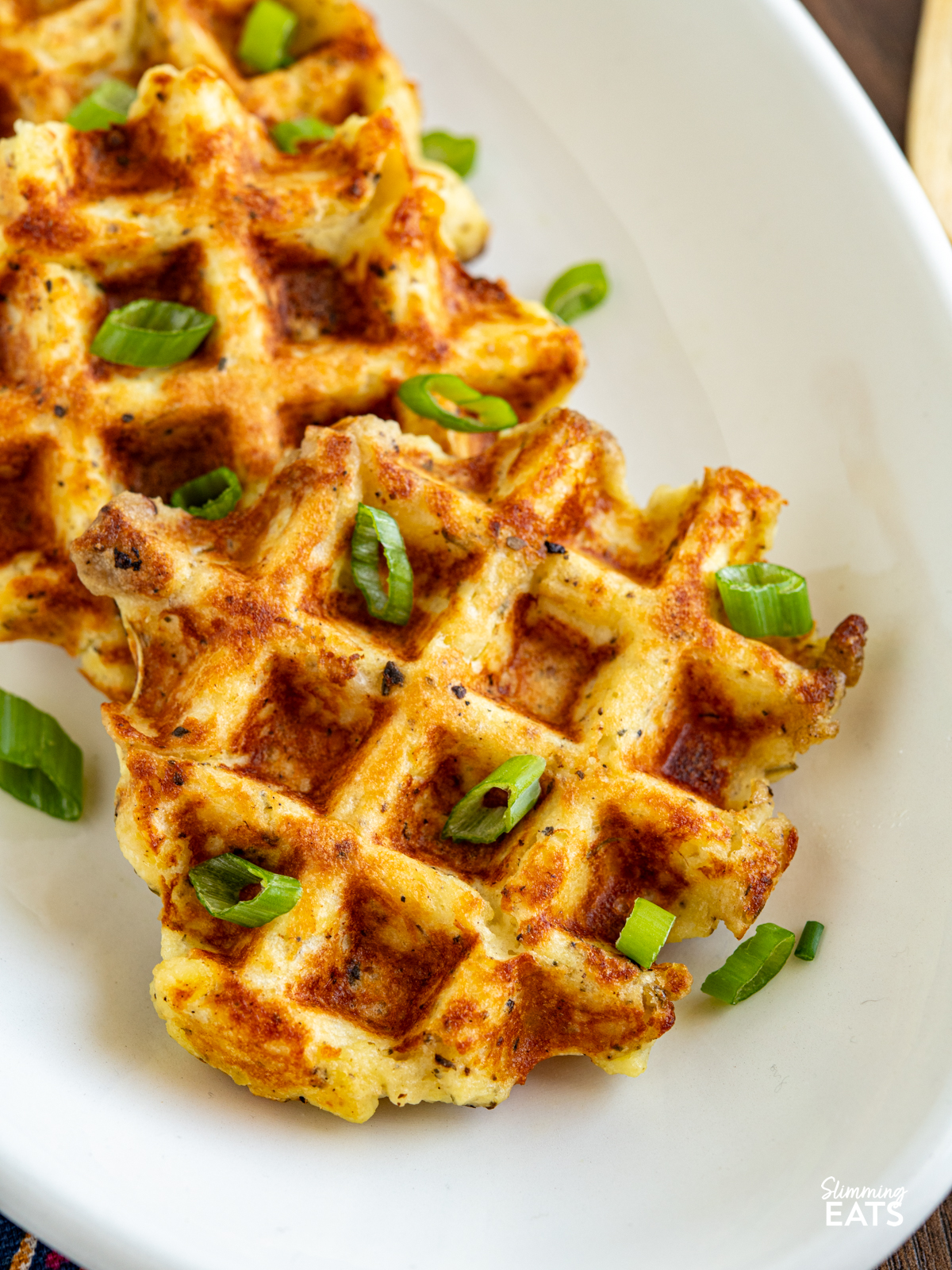 close up cooked potato waffles on a white oval plate garnished with chopped spring onion