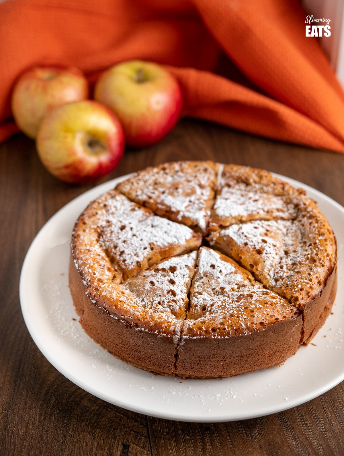 apple sultana cake on white plate with scattered apples and orange cloth in background