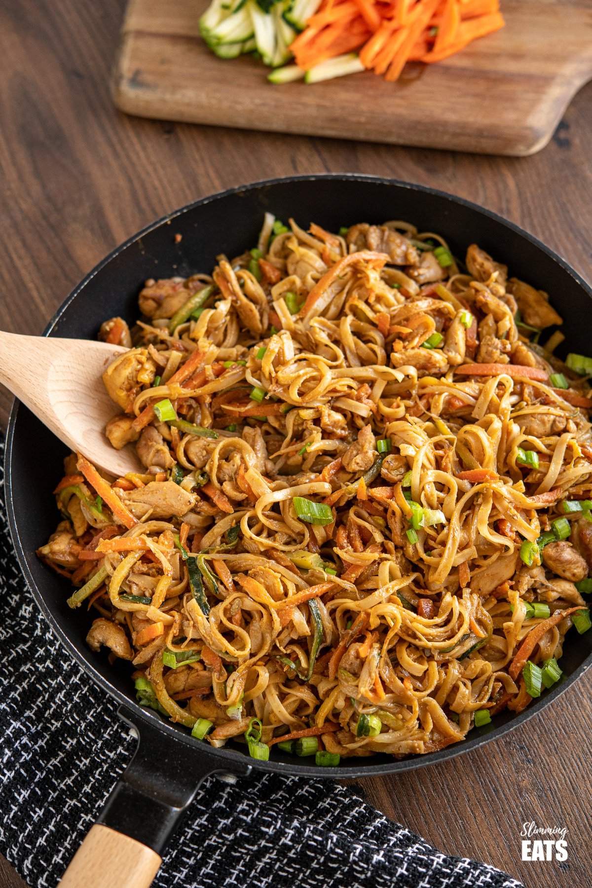 Easy Peanut Chicken Noodles in frying pan with wooden spoon, chopping board with carrots and zucchini in background