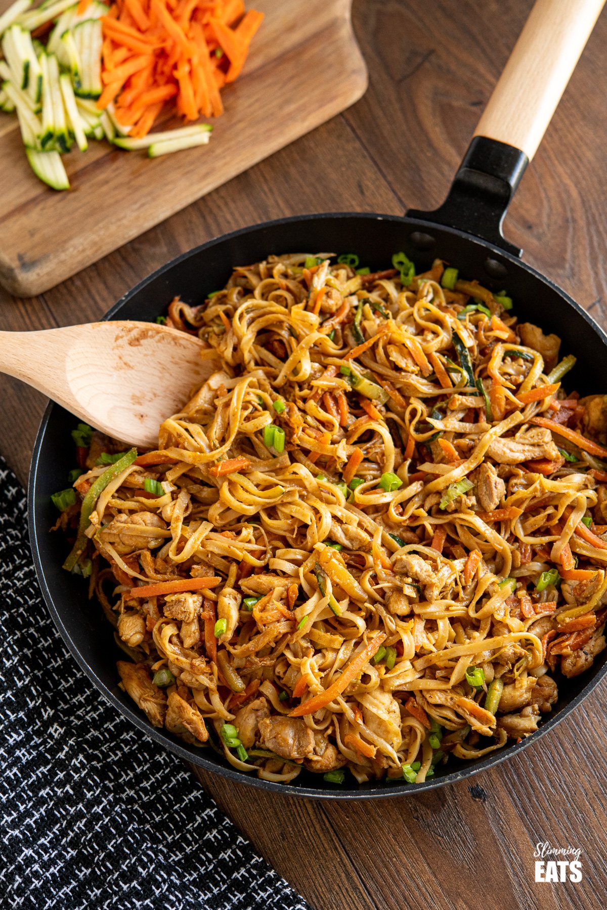 Easy Peanut Chicken Noodles in black frying pan with wooden handle, chopped board with carrots and zucchini in background