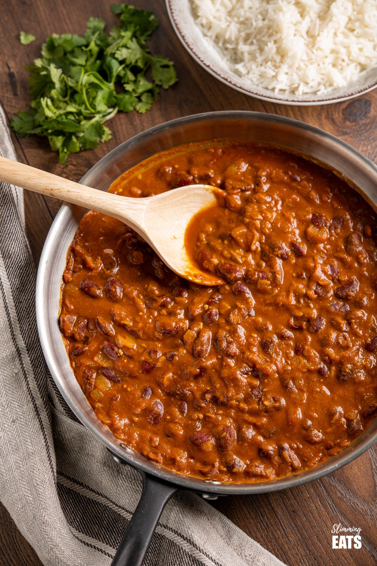 Kidney Bean Curry (Rajma) in stainless steel frying pan with wooden spoon and bowl or rice and cilantro in background